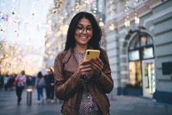 Low angle of cheerful ethnic young female in casual outfit wearing glasses looking at screen of smartphone and messaging while standing in city street on sunset