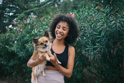 Cheerful youthful African American woman in casual wear hugging cute Chihuahua while standing in lush green city park and smiling
