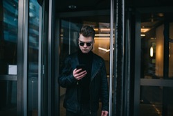 Concentrated young masculine stylish man in black coat and round eyeglasses standing in front of revolving door of office building and using smartphone for checking messages