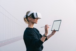 Young female extend hand with digital pencil for interactive touch over air while standing in against bridge construction wearing virtual reality headset. Hipster girl designing pen copy space with VR