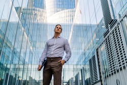 Low angle of youthful male entrepreneur in formal clothes with hand in pocket standing between modern buildings with glass walls and looking away with tablet in hand