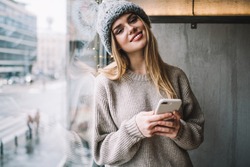 Half length portrait of millennial blogger with modern cellular gadget smiling at camera, happy female generation Z dressed in knitted pullover and winter hat connecting to 4g wireless on mobile