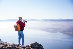 Back view of full body of female photographer with backpack and photo camera taking picture of amazing scenery with foggy landscape and river in sunny day