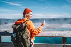 Smiling female tourist with big backpack standing near hot spring in Yellowstone National Park and doing video call on mobile phone