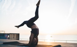 Side view of young woman in bodysuit practicing yoga in Supported Headstand with split while training on seaside at sunrise