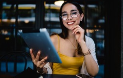 Portrait of cheerful Caucasian female blogger in eyewear smiling at camera during time for online networking via digital tablet, joyful hipster girl with modern touch pad posing in street cafe