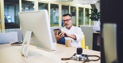Middle aged male manager watching uploaded photos on tablet sitting at desk with computer photo camera and mug with coffee on break