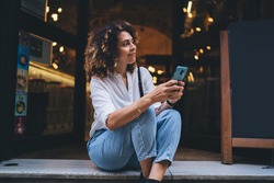 Pretty Caucaisan woman with modern cellphone device in hands resting at touristic street sitting outdoors and smiling, charming hipster girl holding smartphone technology for network and looking away