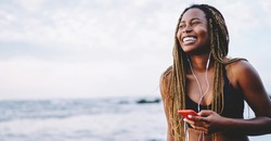 Cheerful African American woman with dreads listening audio songs from cell player, happy Dark skinned jogger in sportive tracksuit enjoying music in earphones connected to cellular application