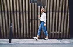 Side view of stylish positive middle aged female in jeans and sneakers strolling along street near modern building and drinking coffee