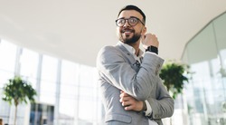 From below of delight male entrepreneur in formal wear and modern eyeglasses standing in office and smiling while looking away