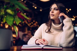 Dreamy female in casual sweater speaking on phone while sitting with notepad and pen at cozy illuminated cafe and looking away