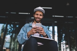 Happy Spanish programmer with closed netbook device using cellphone technology in city, portrait of cheerful hipster guy holding smartphone and laptop and smiling at camera while resting at stairs