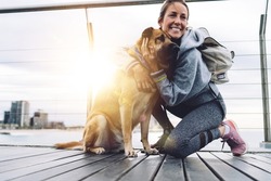 Happy Caucasian woman dressed in sportive clothing resting at boardwalk pier hugging and stroke lovely pet, fit girl with backpack smiling on leisure, concept of human love and friendship to dogs