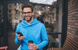 Half length of funny hipster guy in classic spectacles holding modern cellphone gadget and takeaway caffeine beverage and smiling at camera during weekend leisure in city, happy male blogger posing