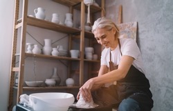 Smiling senior caucasian female master molding clay pot on pottery wheel in art studio. Small business and entrepreneurship. Home hobby, entertainment and leisure. Woman wearing work apron. Sunny day