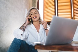 Portrait of joyful hipster girl smiling at camera during positive cell communication via smartphone application, cheerful female millennial with laptop computer posing while making friendly calling
