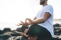 Concentrated male yogi dressed in comfortable wear meditating in lotus pose enjoying relaxation and concentration time at seashore rock, concept of holistic recovery and mental healthy lifestyle