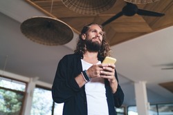 Contemplative bearded male blogger with modern cellular device thinking about web content idea, pondering Caucaisan man with long hair using mobile gadget during leisure for pda communication