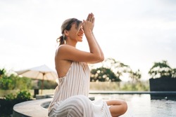 Young joyful woman practicing yoga and meditating on swimming pool edge in yard of resort hotel. Tourism, vacation and weekend. Caucasian girl. Sunset. Idyllic and tranquility lifestyle on Bali island