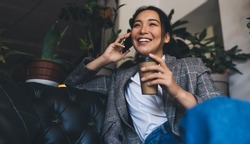 Happy Asian female in smart casual jacket enjoying coffee break for calling and talking via smartphone technology, cheerful woman with takeaway caffeine beverage phoning via mobile application