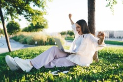 Side view of thrilled young female sitting on grass with raised arms and closed fists while looking at screen of laptop during remote work on project