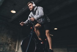 Low angle of young male in headphones looking away while sitting on spinning stationary cycle and listening to music in wireless headphones while doing cardio workout in gym