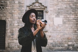 Pensive black female tourist in casual clothes and hat standing on Barcelona street and taking pictures on photo camera during vacation