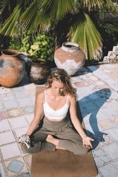 High angle of young female sitting with legs crossed on mat on floor and meditating with prayer beads in hand in daylight near green palms