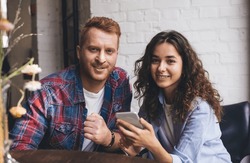 Portrait of Caucasian male and female with cellular gadget looking at camera during leisure time, millennial best friends with smartphone technology posing during date meeting in cafe interior