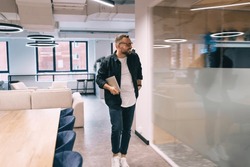 Positive man in smart casual clothes and glasses walking near glass wall with laptop while working in spacious modern office