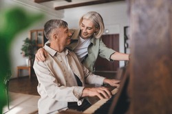 Happy aged female in casual clothes hugging man playing melody on piano during weekend together at home looking at each other and smiling
