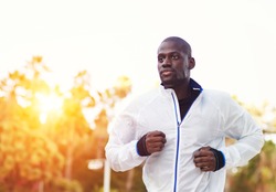 Young fit man in white windbreaker running at orange sunset outdoors, autumn running, attractive dark skinned runner jogging fast in the park with beautiful sunset light on background, sport concept