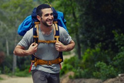Portrait of a young smiling wanderer man with backpack goes on up the road with copy space, happy hiker walking lonely in mountains at summer day, traveler enjoying his recreation time in nature