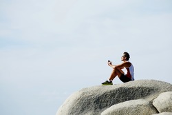 Male runner listening to music in headphones while sitting on rock cliff against sky copy space background for your text message or advertising, weary mature jogger resting after workout jog outdoors