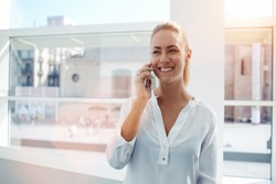 Smiling businesswoman dressed in formal wear having pleasant conversation on mobile phone with friend, happy attractive female speaking on cell telephone while resting in  office interior after work