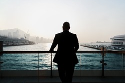 Back view of a young confident man financier is thinking about something, while is standing outdoors against sea port. Silhouette of male intelligent managing director is resting after hard work day