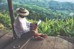 Man traveler is using digital tablet, while is sitting against beautiful Asian scenery during summer journey. Male wanderer is holding touch pad, while is relaxing outdoors during his trip in Thailand