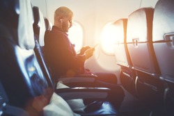 Man entrepreneur is watching video on mobile phone, while is sitting in plane near window with sun rays during his business trip. Hipster guy is listening to music in headphones via cell telephone