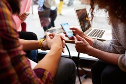 Closely image of woman is making an on-line purchase via cell telephone, while is sitting with friends in hipster cafe interior. Young female is reading information on web page via her mobile phone