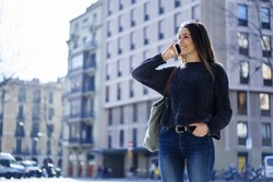 Smiling trendy dressed female tourist standing on promotional background satisfied with connection in roaming talking on mobile, attractive hipster girl in stylish wear having phone conversation