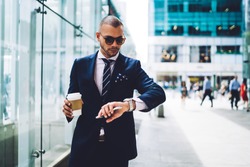 Confident male entrepreneur  checking time on watch during break while hurrying for meeting, serious businessman in formal suit standing on urban settings with coffee takeaway looking at wristwatch