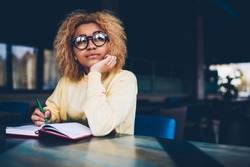 Thoughtful young woman in eyeglasses writing to do list of goals writing in diary in coffee shop, female student dreaming about weekends while making notes in personal organizer planning day
