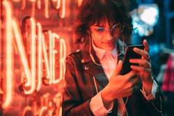 Young stylish female hipster standing near neon red light in night city and listening to music in headphones on modern smartphone. Woman watching media video on cellphone device