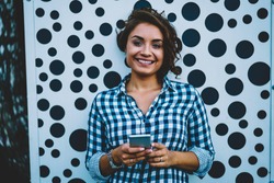 Half length portrait of cheerful female internet user doing shopping online and installing mobile aplication with webstore on modern telephone standing outdoors against promotion background