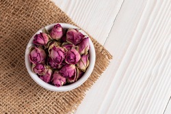 Dried damask rose buds in small bowl on white wooden table. Top view.
