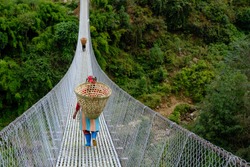 Nepalese woman carrying a wicker basket on her back, walking on a hanging bridge. Nepal, Himalaya. During trekking on trail from Ghangaruk to Phedi