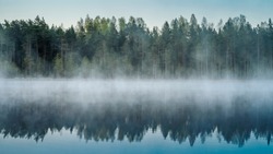 Cold summer morning in the forest with lake, forest reflection and mist on the water surface.