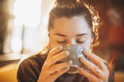 portrait of a beautiful woman drinking coffee or tea with a large mug sitting in a room where a lot of bright sunlight and sunlight go through the windows and create a warm light atmosphere