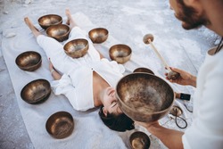man conducts therapy Tibetan singing bowls for a girl lying on the ground in the desert surrounded by copper bowls